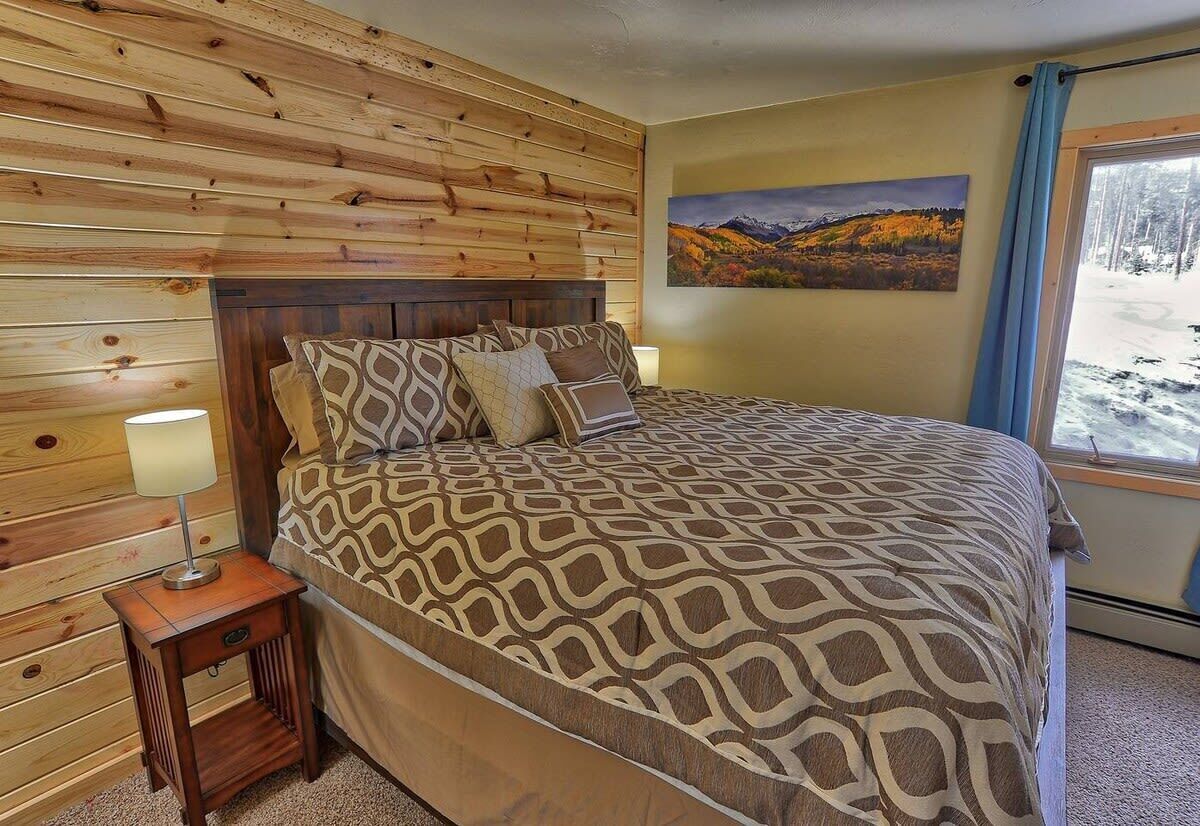 A bedroom with a wooden headboard, patterned bedding, a bedside table with a lamp, and a window with a view of a snowy landscape.
