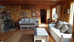 A cozy cabin interior with a stone fireplace, a gray sofa with compass-themed pillows, a wooden coffee table, and a lamp on a side table. - The Haystack House-Cannon Beach Oceanfront Home Steps to Haystack Rock & Midtown (Cannon Beach)