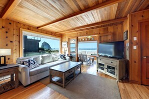 A wooden cabin interior with a sofa, coffee table, TV, and a view of the ocean. - The Haystack House-Cannon Beach Oceanfront Home Steps to Haystack Rock & Midtown (Cannon Beach)