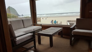 A room with a sofa, a table, and a chair, overlooking a beach with people and a rock formation. - The Haystack House-Cannon Beach Oceanfront Home Steps to Haystack Rock & Midtown (Cannon Beach)