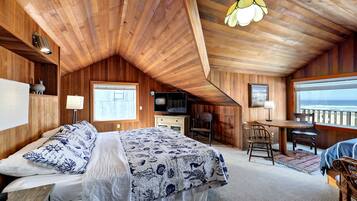 A cozy room with a sloped ceiling, wooden walls, a bed with a patterned bedspread, a desk with a chair, a television, and a window overlooking a beach.