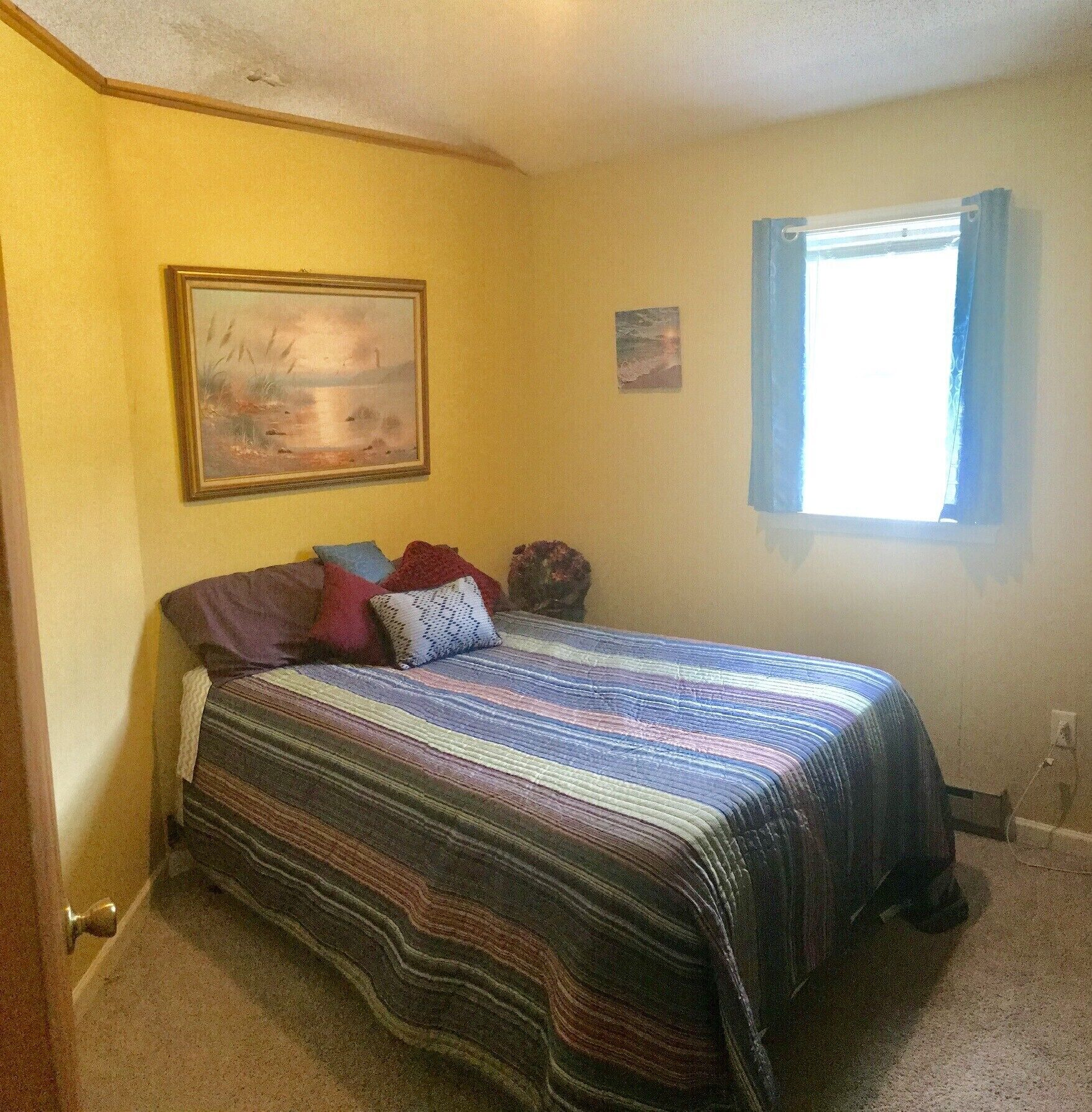 A bedroom with a bed, a painting on the wall, a window with curtains, and a carpeted floor.