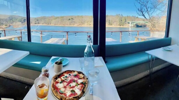 Een eethoek met een tafel gedekt voor twee personen, pizza, een fles en glazen, met uitzicht op een waterlichaam met een brug in de verte.