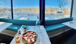 A dining area with a table set for two, a pizza, a bottle, and glasses, overlooking a body of water with a bridge in the distance. - Honesdale Escape ~ A Place to Remember. (Honesdale)
