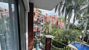 A balcony with a view of a swimming pool, palm trees, and a building with red brick and white trim. - BEST DEAL IN HEART OF TAMARINDO. Block from the beach, walk to everything! (Tamarindo)