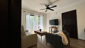 A living room with a ceiling fan, a TV on a wooden stand, a sofa, a chair, and a large window with sheer curtains. - BEST DEAL IN HEART OF TAMARINDO. Block from the beach, walk to everything! (Tamarindo)