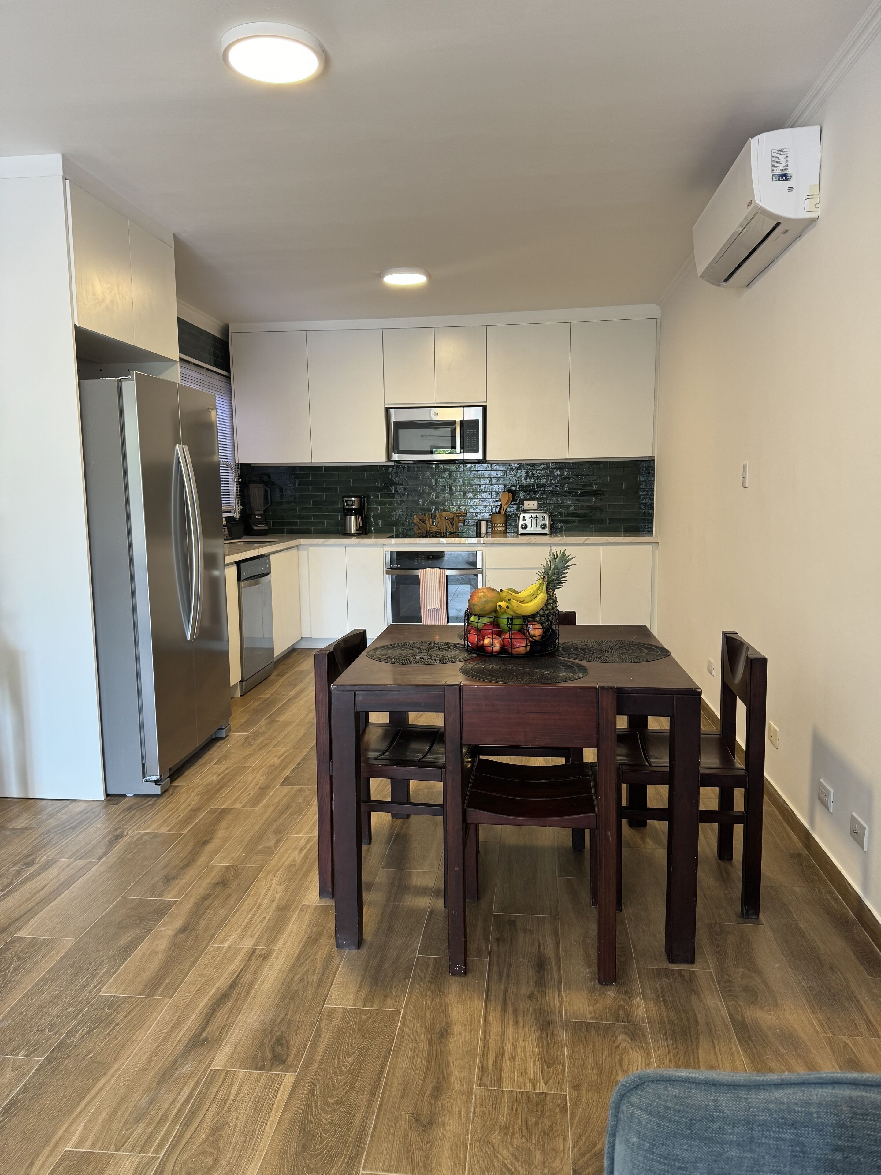 A modern kitchen with white cabinets, a stainless steel refrigerator, a microwave, and a fruit basket on the dining table.