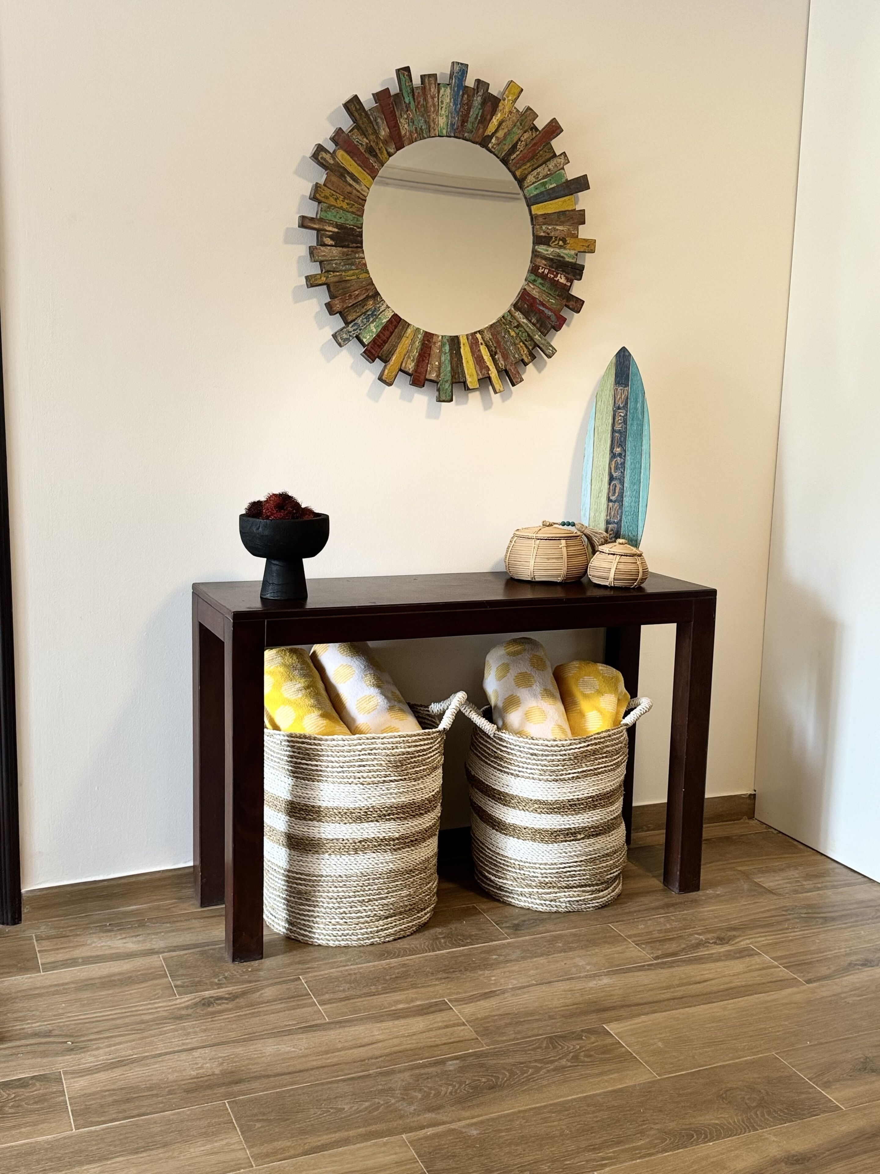 A wooden console table with two woven baskets underneath, a mirror with a wooden frame, and a surfboard with a 'WELCOME' sign.