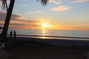 Escena de playa con un atardecer sobre el océano, palmeras y algunas personas de pie en la orilla.