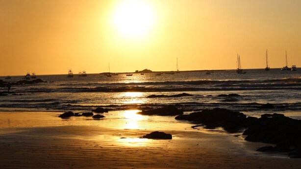A beach scene at sunset with boats in the water and rocks on the shore. - BEST DEAL IN HEART OF TAMARINDO. Block from the beach, walk to everything! (Tamarindo)