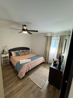 Un dormitorio con cama, mesita de noche, escritorio, ventilador de techo y ventana con cortinas.
