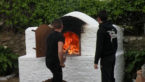 A traditional wood-fired oven with two individuals nearby, surrounded by greenery. - Pizania (Kalymnos)