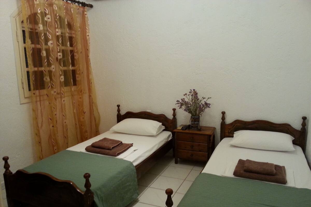 A room with two wooden beds, each with a green bedspread and a white pillow. There is a wooden nightstand between the beds with a vase of flowers.