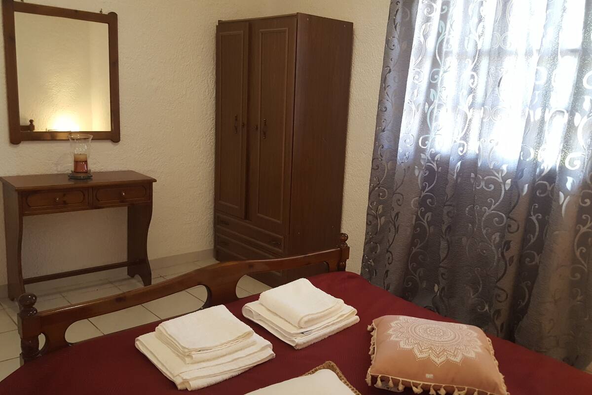 A bedroom with a wooden wardrobe, a bedside table with a lamp, a bed with neatly folded white towels, and a patterned curtain.