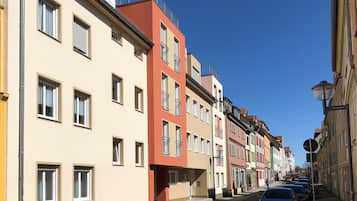 Une rangée d’immeubles à étages avec des balcons, un ciel dégagé et des voitures garées le long de la rue.