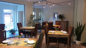 A dining area with wooden tables and chairs, a glass partition, and potted plants. - Gästehaus Luise (Gotha)