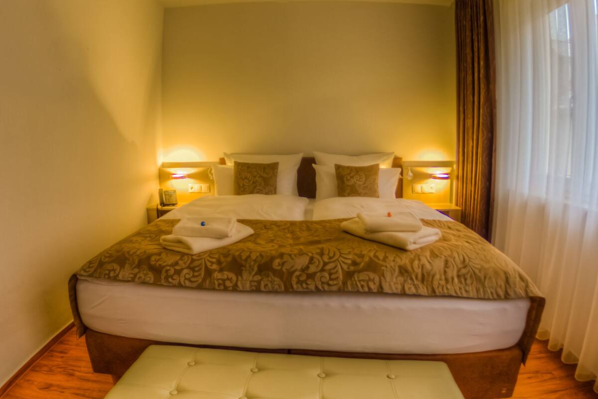 A hotel room with a large bed, two bedside lamps, a bedside table, and a window with curtains.