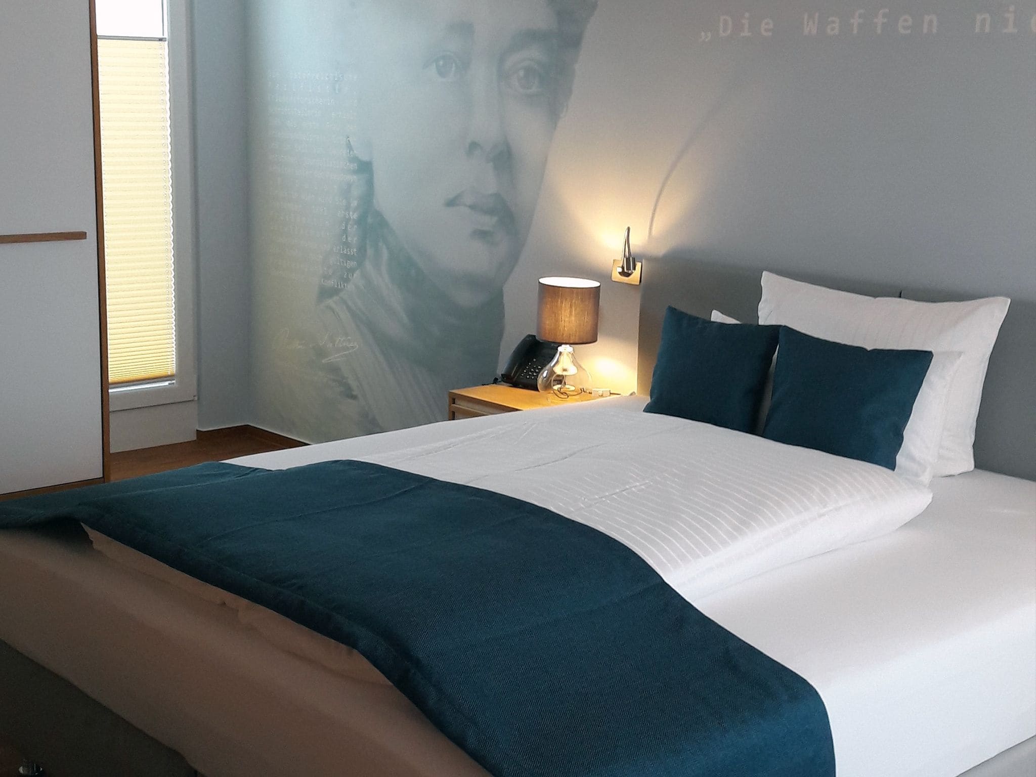 A neatly made bed with a white and teal bedspread, a bedside lamp, and a wall with a partial view of a person's face.