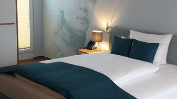 A neatly made bed with a white and teal bedspread, a bedside lamp, and a wall with a partial view of a person's face.