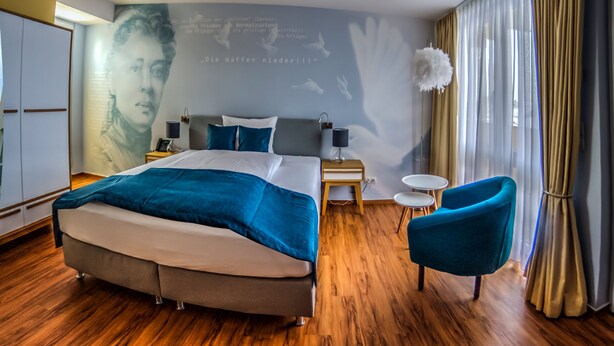 A modern hotel room with a large bed, a small side table, a chair, and a wall with a mural. - Gästehaus Luise (Gotha)