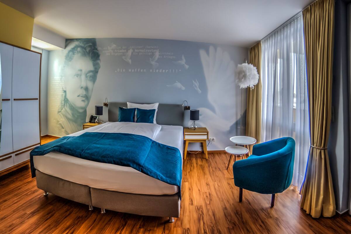 A modern hotel room with a large bed, a small side table, a chair, and a wall with a mural.