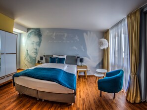 Una habitación de hotel moderna con una cama grande, una mesita auxiliar pequeña, una silla y una pared con un mural.