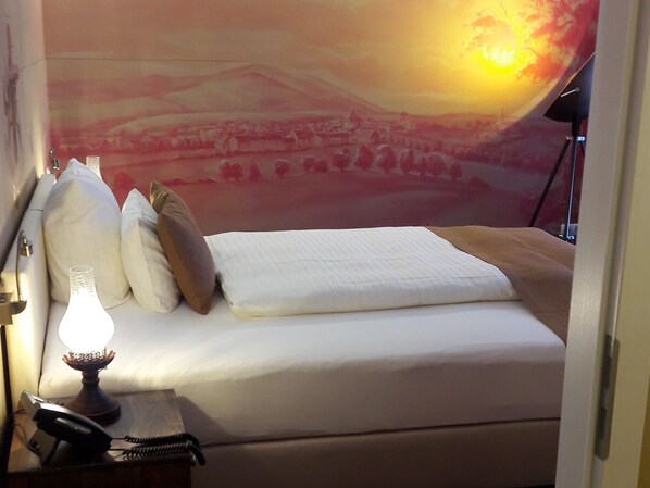 Habitación de hotel con cama, almohadas, lámpara y un mural en la pared que muestra un atardecer sobre un paisaje.