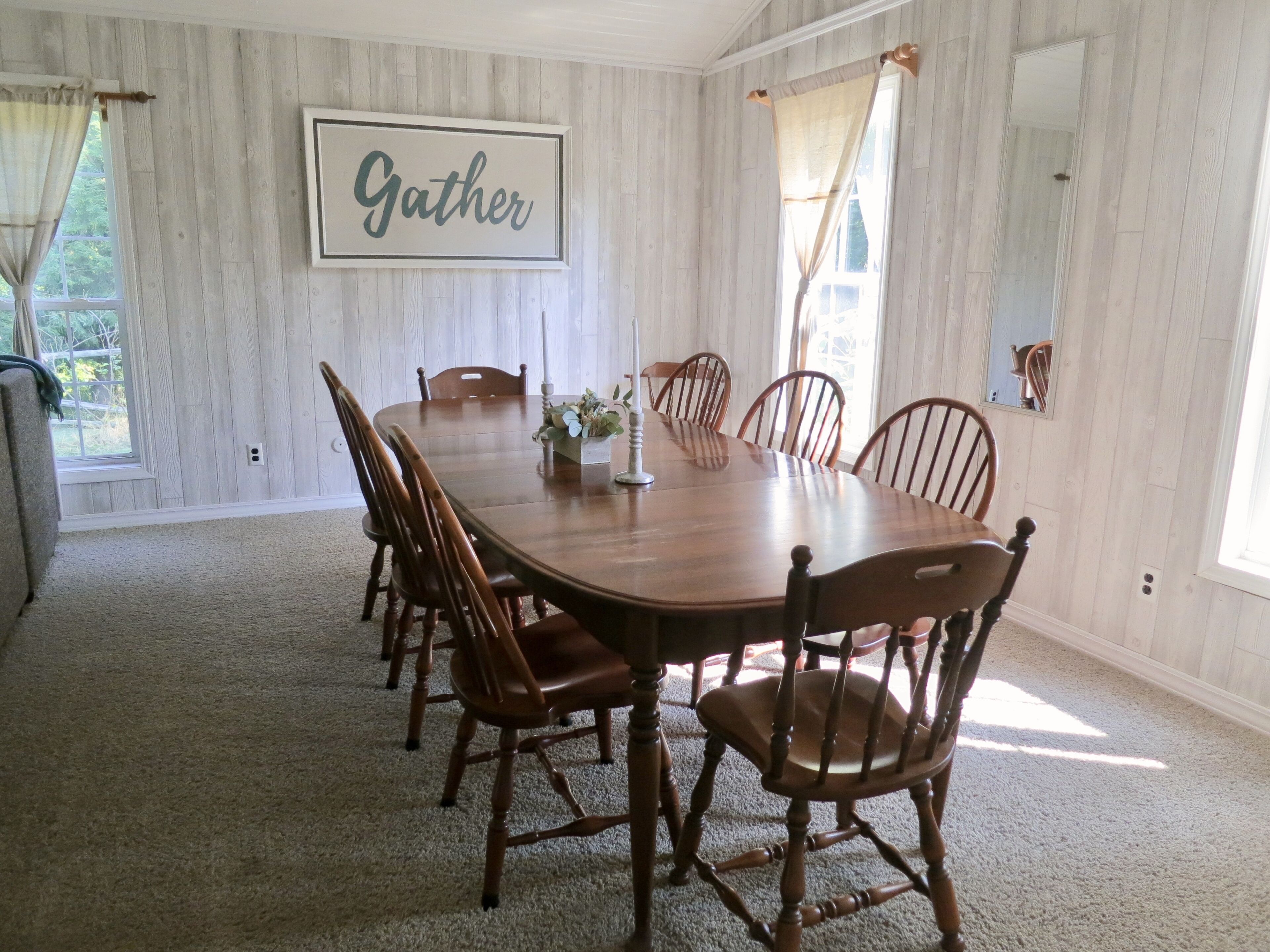 Une salle à manger avec une table et des chaises en bois, un panneau décoratif portant l’inscription « Gather » au mur, et une fenêtre avec des rideaux.