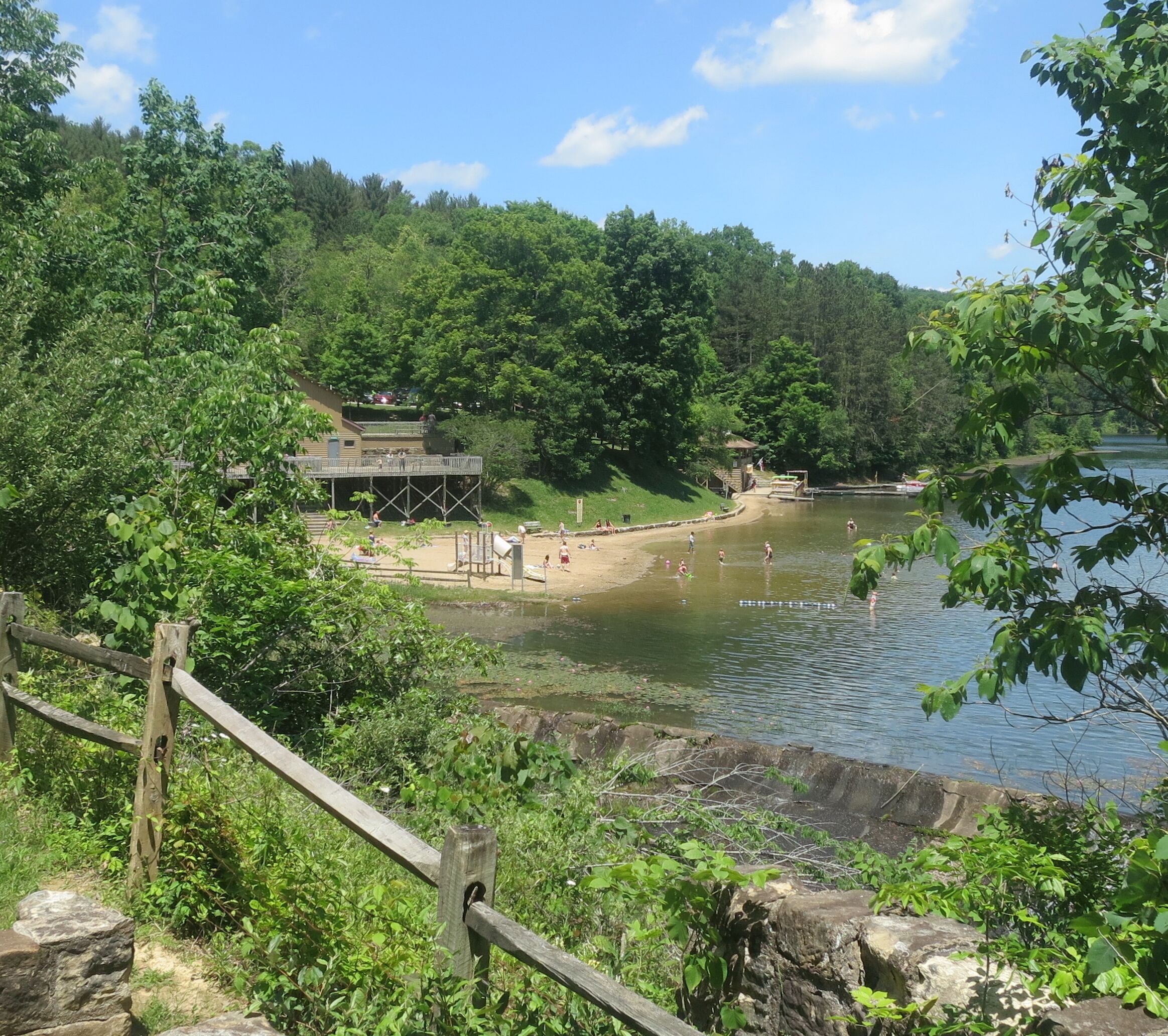 Ein Park am Seeufer mit einer Holzterrasse, einem kleinen Gebäude und einem Sandstrand.
