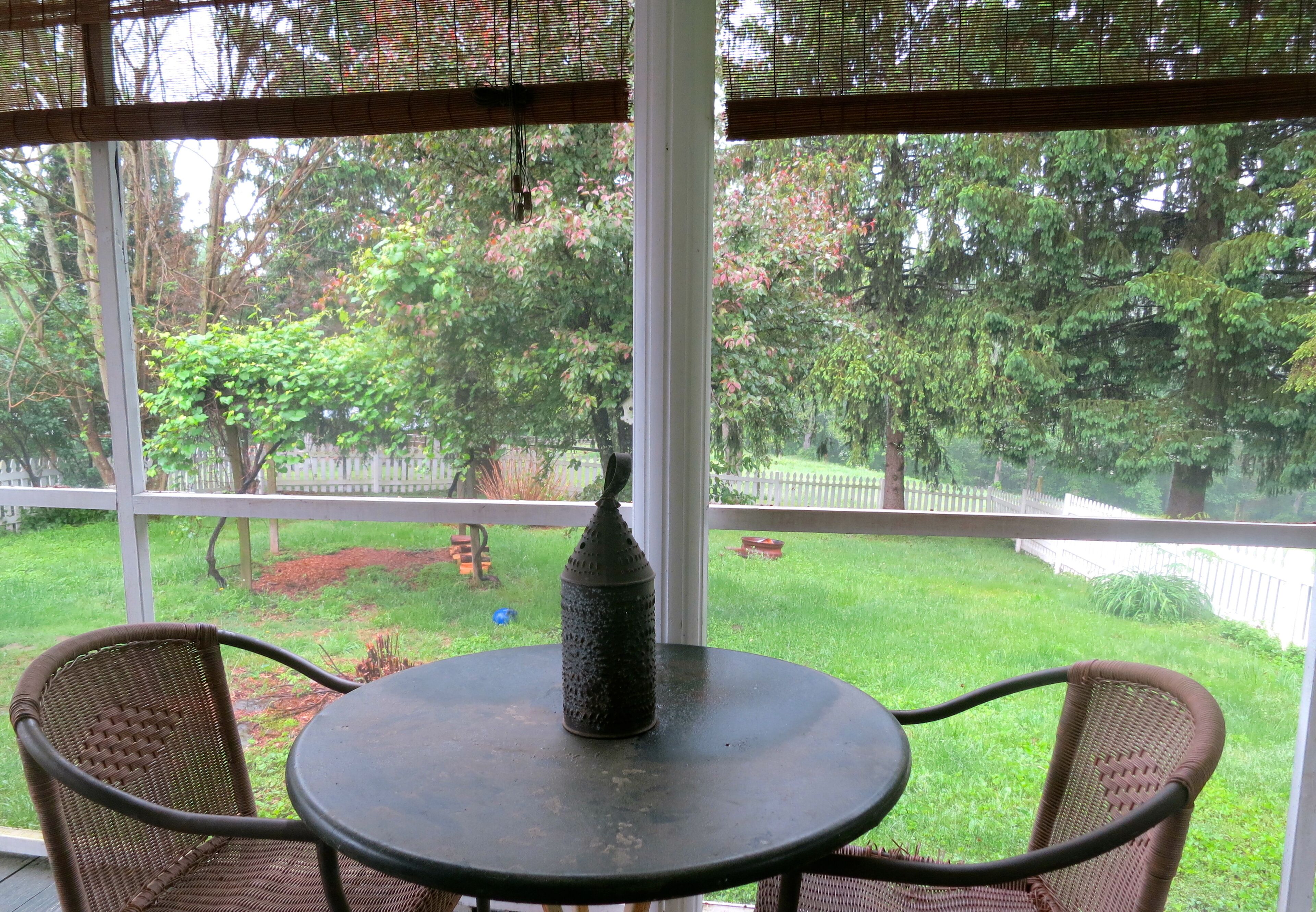 Une terrasse couverte avec une table et des chaises, une bouteille décorative, et une vue sur un jardin luxuriant et une clôture blanche en bois.
