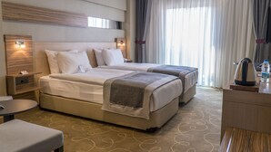 A hotel room with two beds, a desk, a chair, a nightstand, and a large window with curtains. - Greymark Hotel (Izmir)