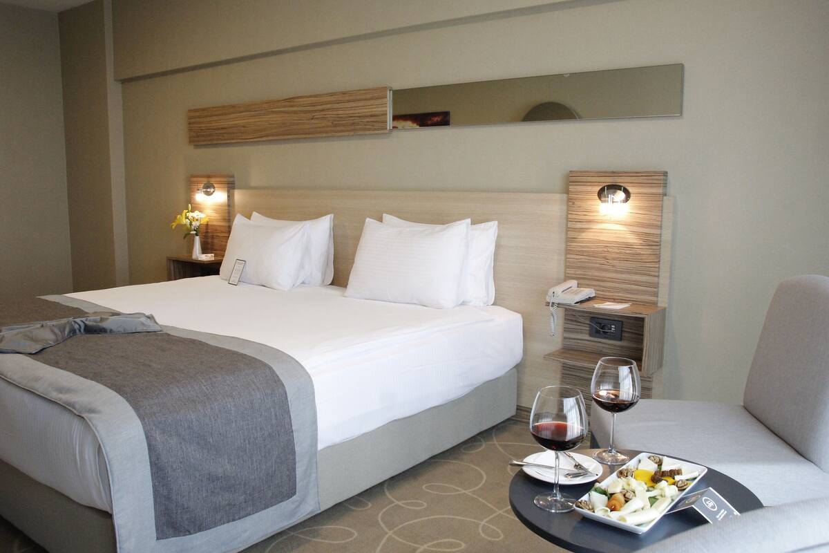 Quarto de hotel com uma cama grande, mesas de cabeceira com luminárias, uma pequena mesa com prato de comida e copos de vinho, e um espelho na parede.