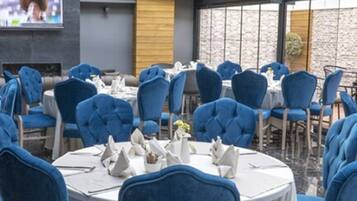 A restaurant with round tables and blue upholstered chairs, a TV screen, and a stone wall in the background.