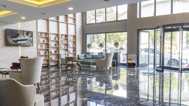 A spacious lobby with a polished marble floor, a seating area with armchairs and a sofa, a bookshelf filled with books, and large windows offering a view of the street outside. - Greymark Hotel (Izmir)