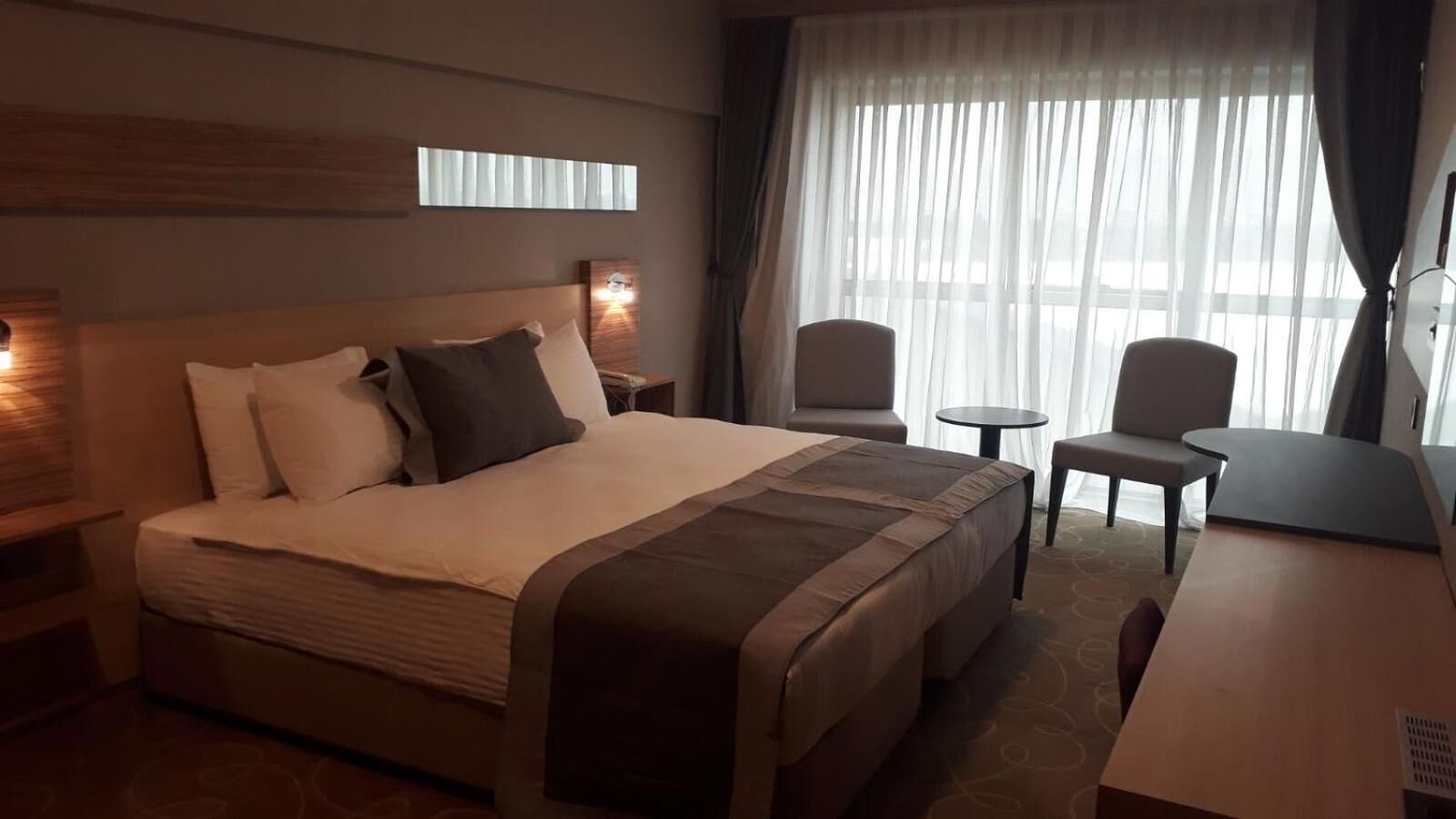 A hotel room with a large bed, two chairs, a small table, and a window with curtains.