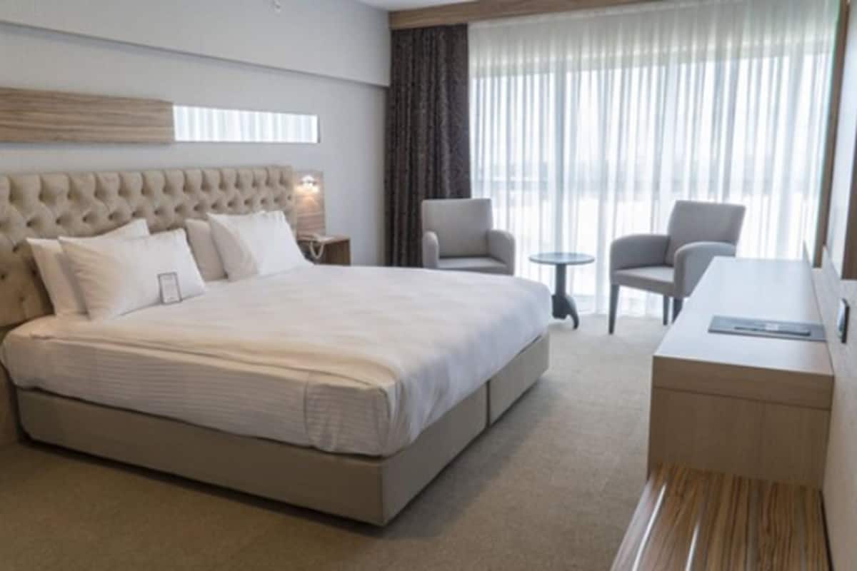 Quarto de hotel com uma cama grande, dois poltronas, uma mesinha e uma escrivaninha com cadeira.
