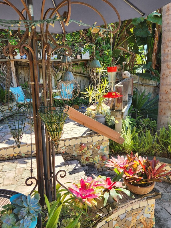 Une terrasse de jardin agrémentée de plantes en pot, d’une chaise en métal et d’un mur décoré de mosaïques.