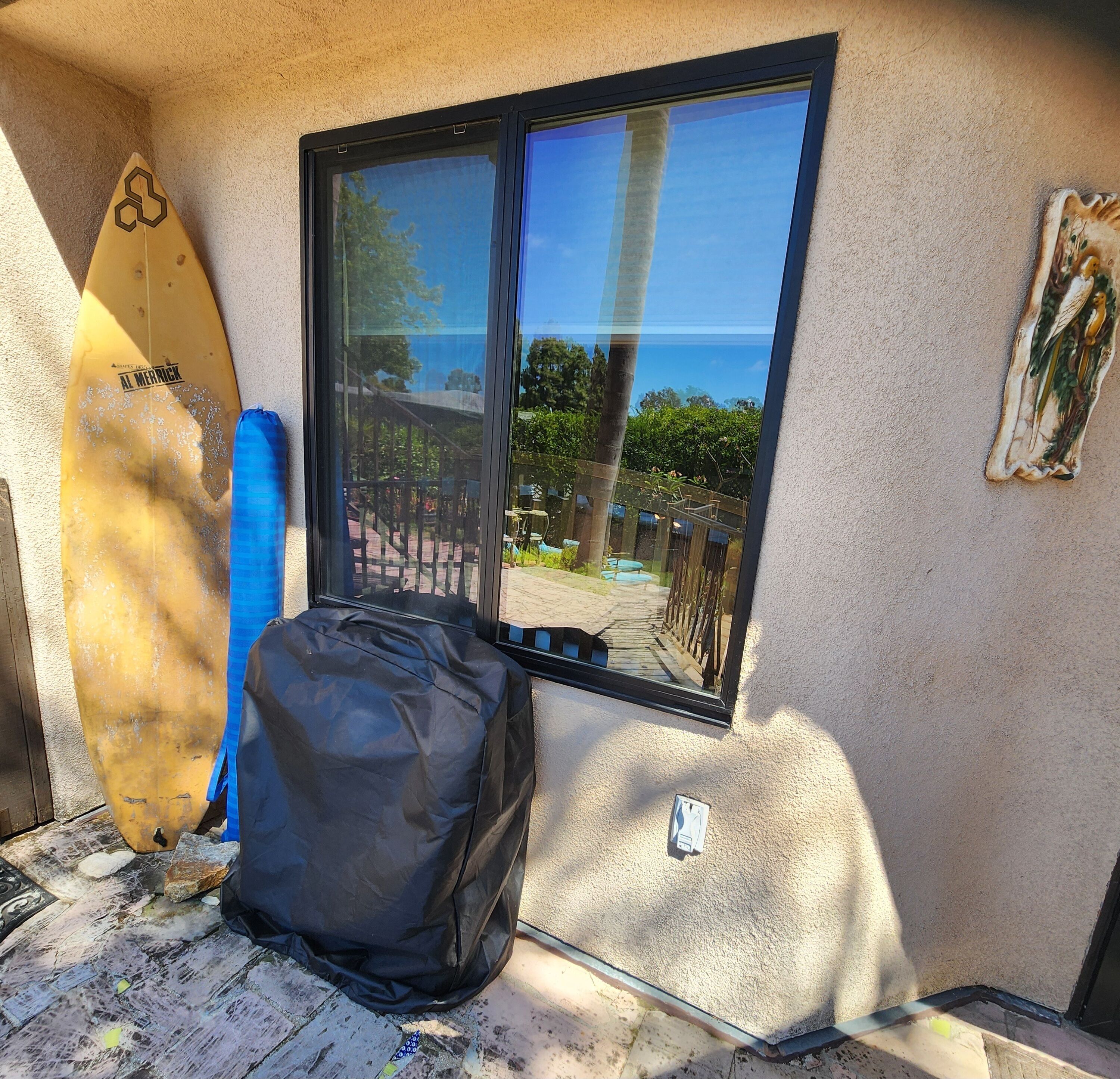 A room with a surfboard leaning against a window, a black bag on the floor, and a small statue on the wall.