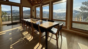 Un comedor con una mesa grande y sillas, ventanas amplias con vistas a la montaña, vigas de madera en el techo y una araña.