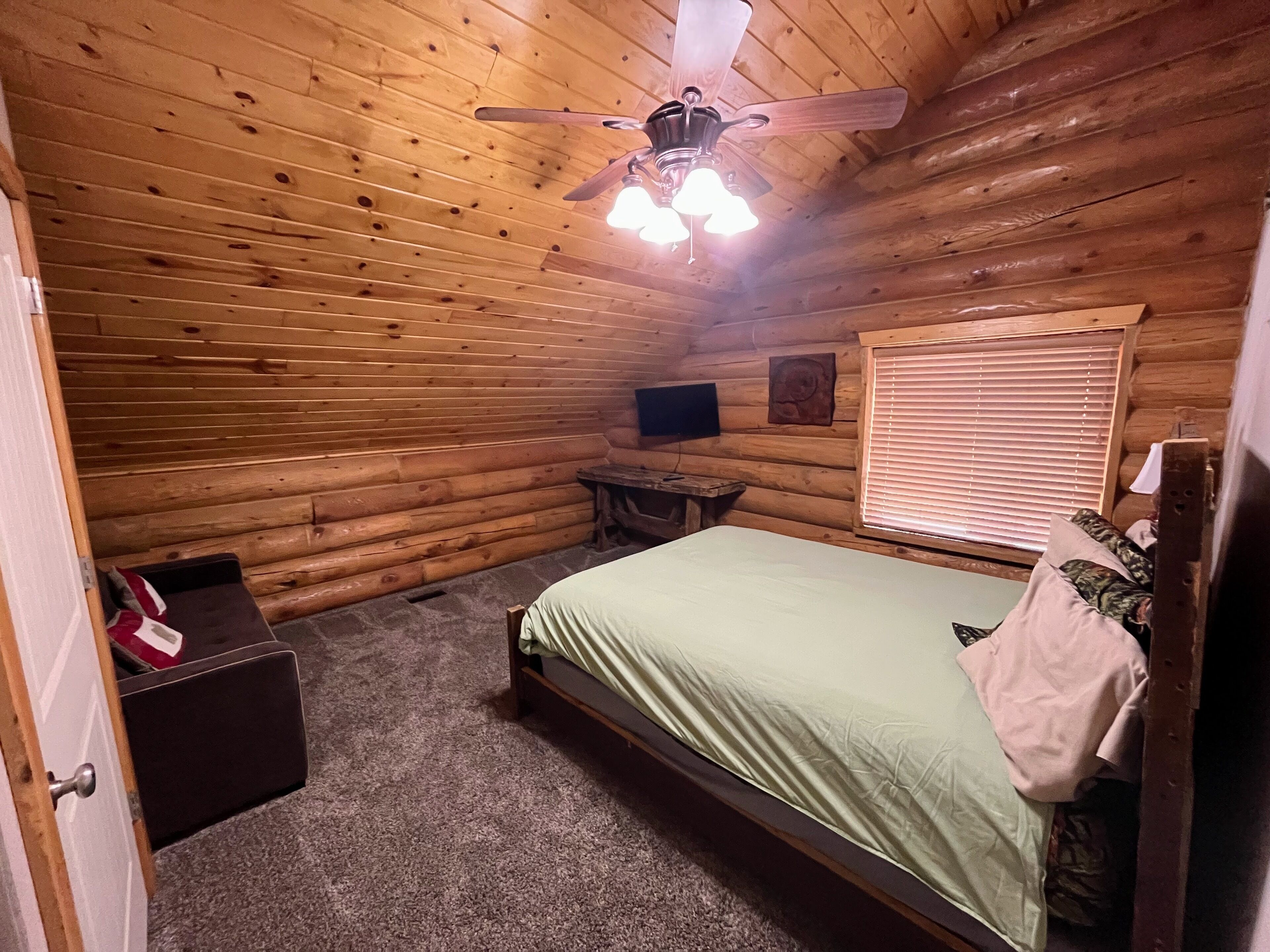 Un dormitorio de cabaña de troncos con una cama, un ventilador de techo, una ventana con persianas, una mesita y un televisor en la pared.