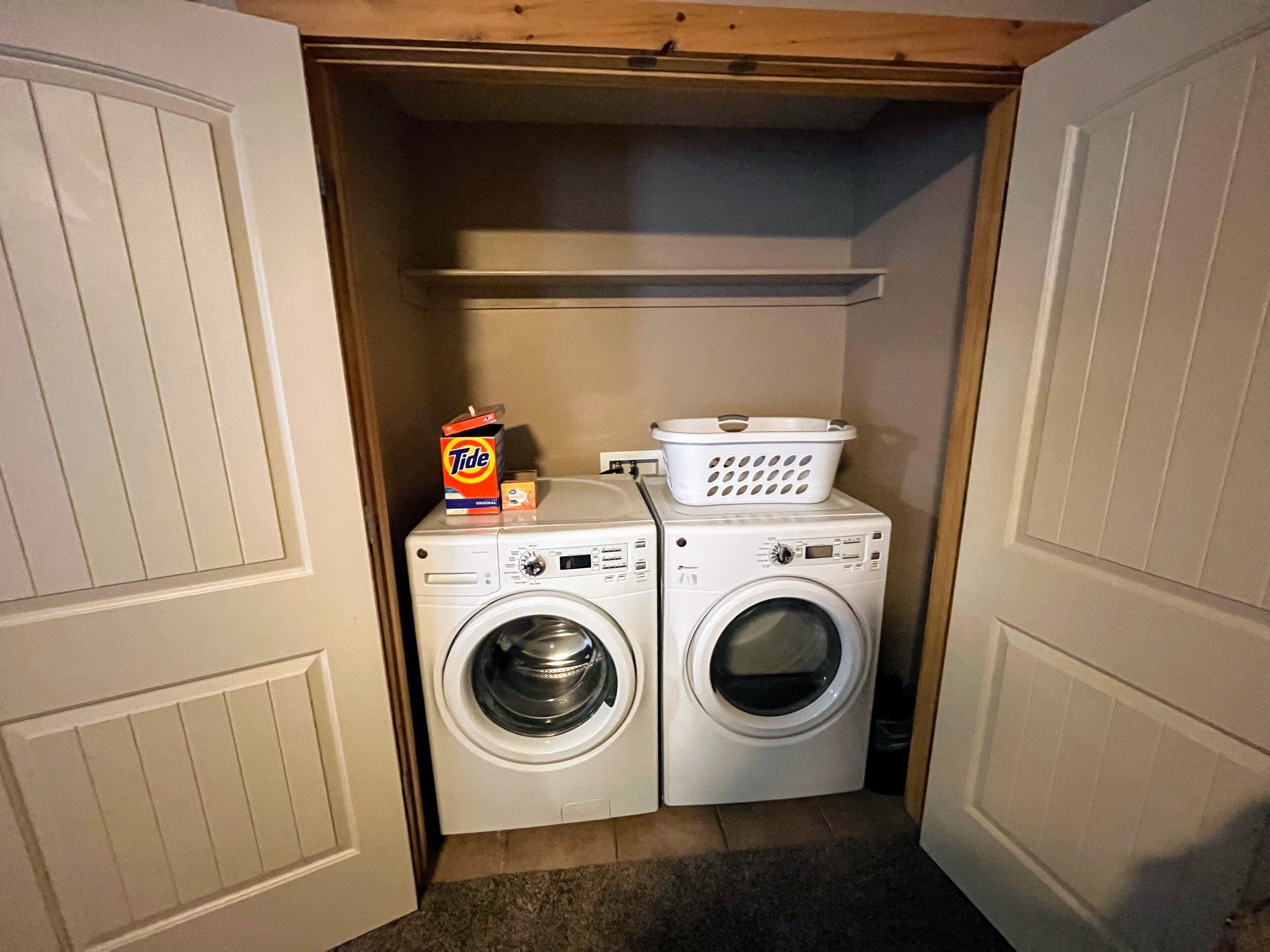 A laundry room with two front-loading washing machines, a Tide detergent box, and a laundry basket.