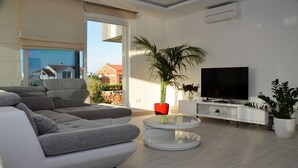 Un salon moderne avec un canapé gris, un téléviseur et une grande fenêtre donnant sur un quartier résidentiel.