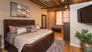 A bedroom with a large bed, a brick wall, a wooden ceiling, and a flat-screen TV.