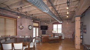 A spacious dining area with a wooden table and chairs, a leather couch, and a television mounted on the brick wall.