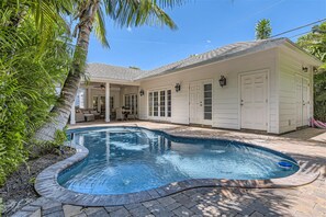 Une piscine à bord incurvé, entourée d’une terrasse dallée. On y trouve également un patio couvert avec des meubles d’extérieur.