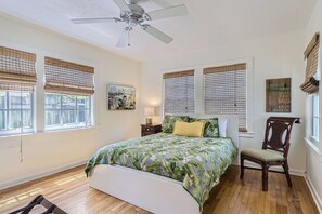 Quarto com cama, uma cadeira, ventilador de teto e janelas com persianas.