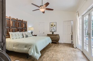 Une chambre à coucher comprenant un lit, une tête de lit en bois, un ventilateur de plafond, une table de chevet, une lampe, une commode, un tableau et une grande fenêtre.
