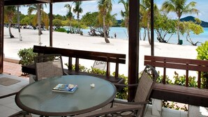 A covered outdoor seating area with a glass table and chairs, overlooking a beach with palm trees and a view of the sea.
