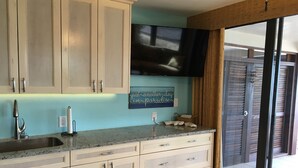 A kitchen with light-colored cabinets, a granite countertop, a stainless steel sink, and a flat-screen TV mounted above the counter.