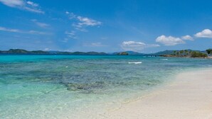 A beach with clear turquoise water, a sandy shore, and distant mountains under a blue sky.